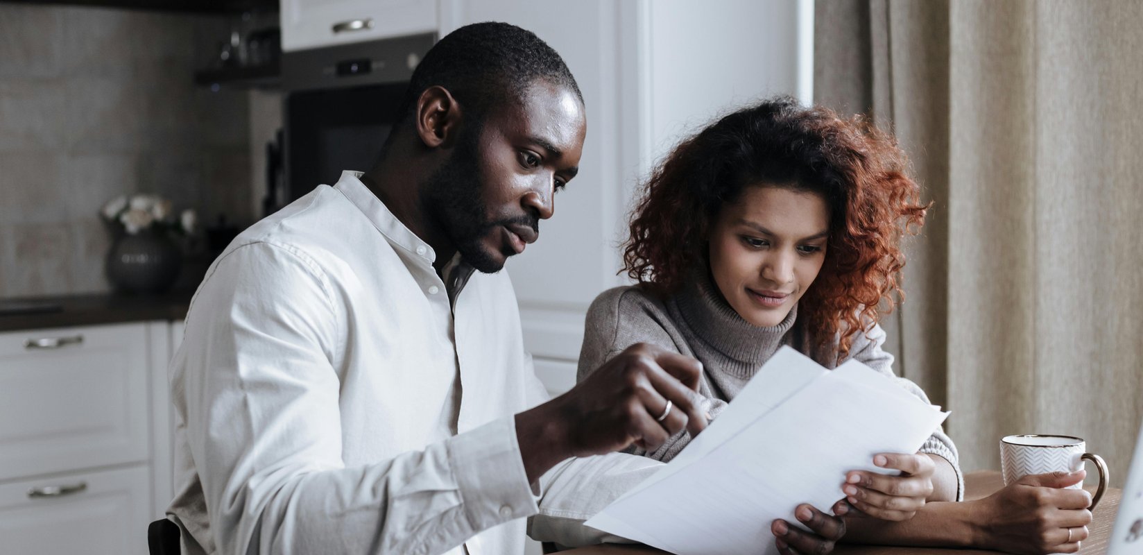A person pointing at a document while a woman holds a coffee cup in a kitchen